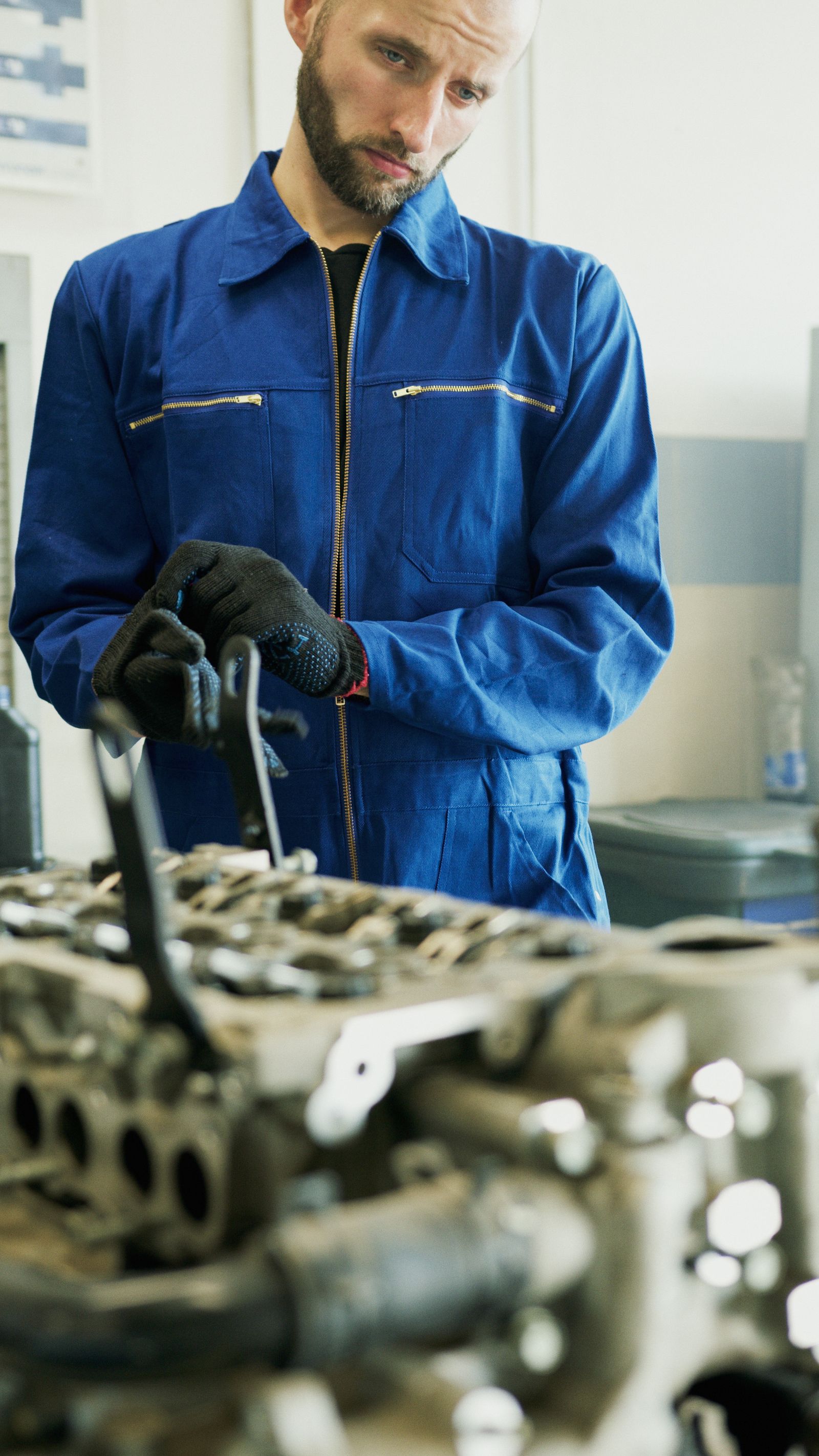 Mechanic wearing blue coveralls and gloves inspecting an engine in a workshop.